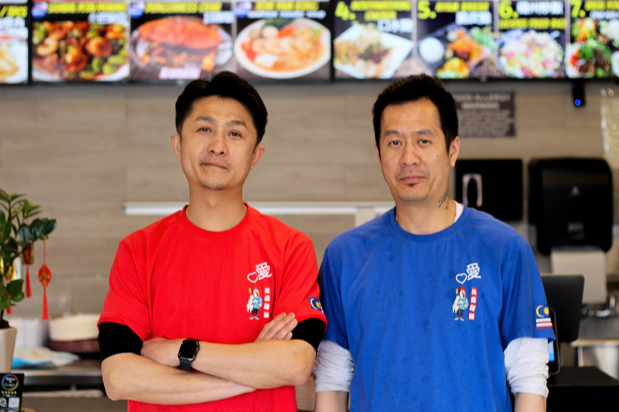 Two brothers, one in a red t-shirt and the other in red, pose for a portrait inside their restaurant.