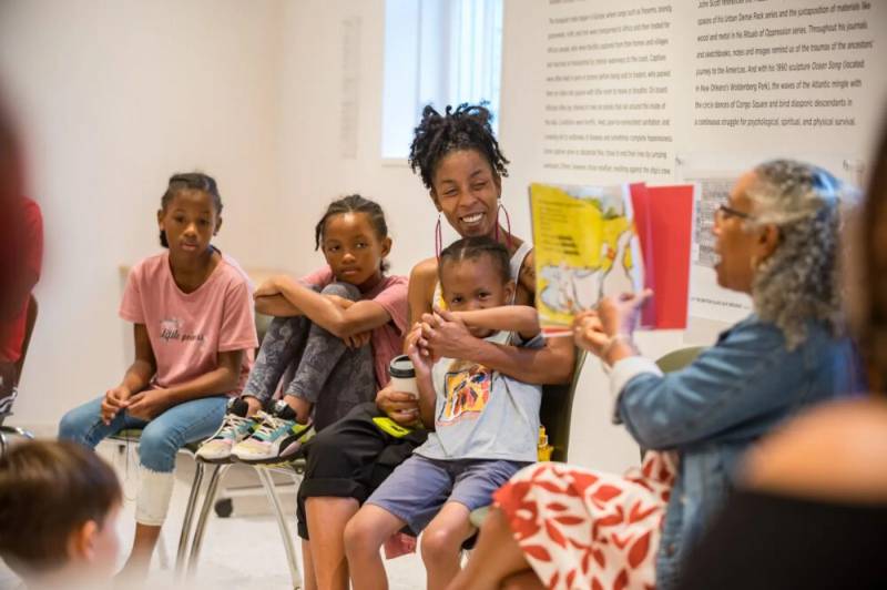three children and adult look on as older adult reads a book