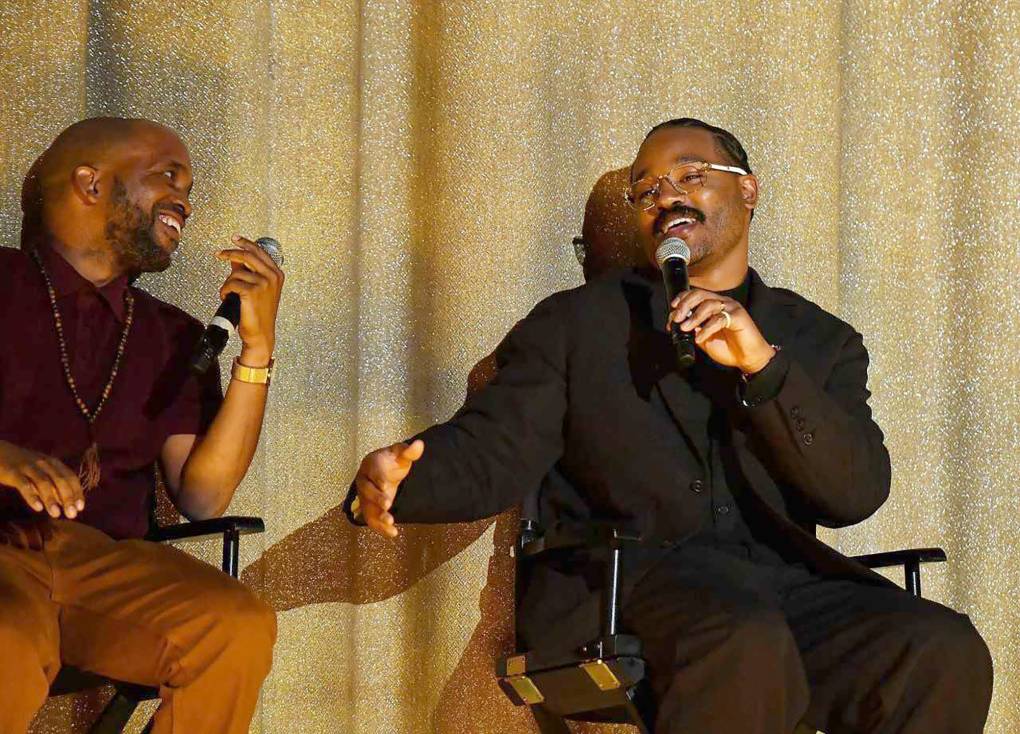 Two African American men sit in directors-style chairs and share conversation while holding microphones in front of a gold curtain