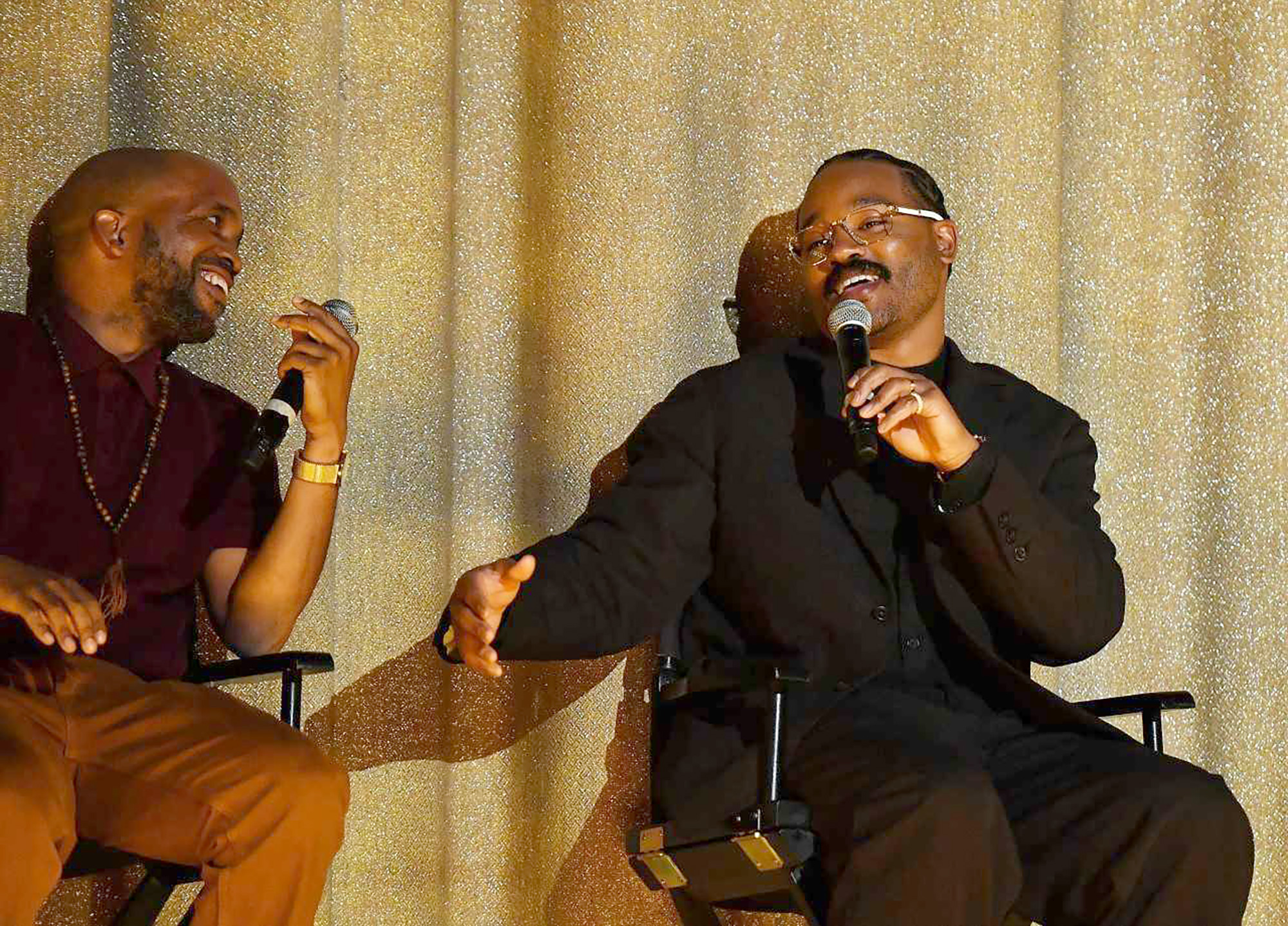 Two African American men sit in directors-style chairs and share conversation while holding microphones in front of a gold curtain