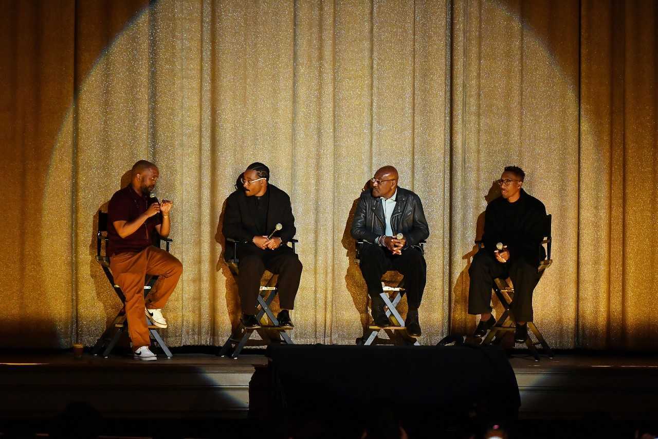 Four Black men sitting onstage in chairs and holding microphones against a gold draped curtain