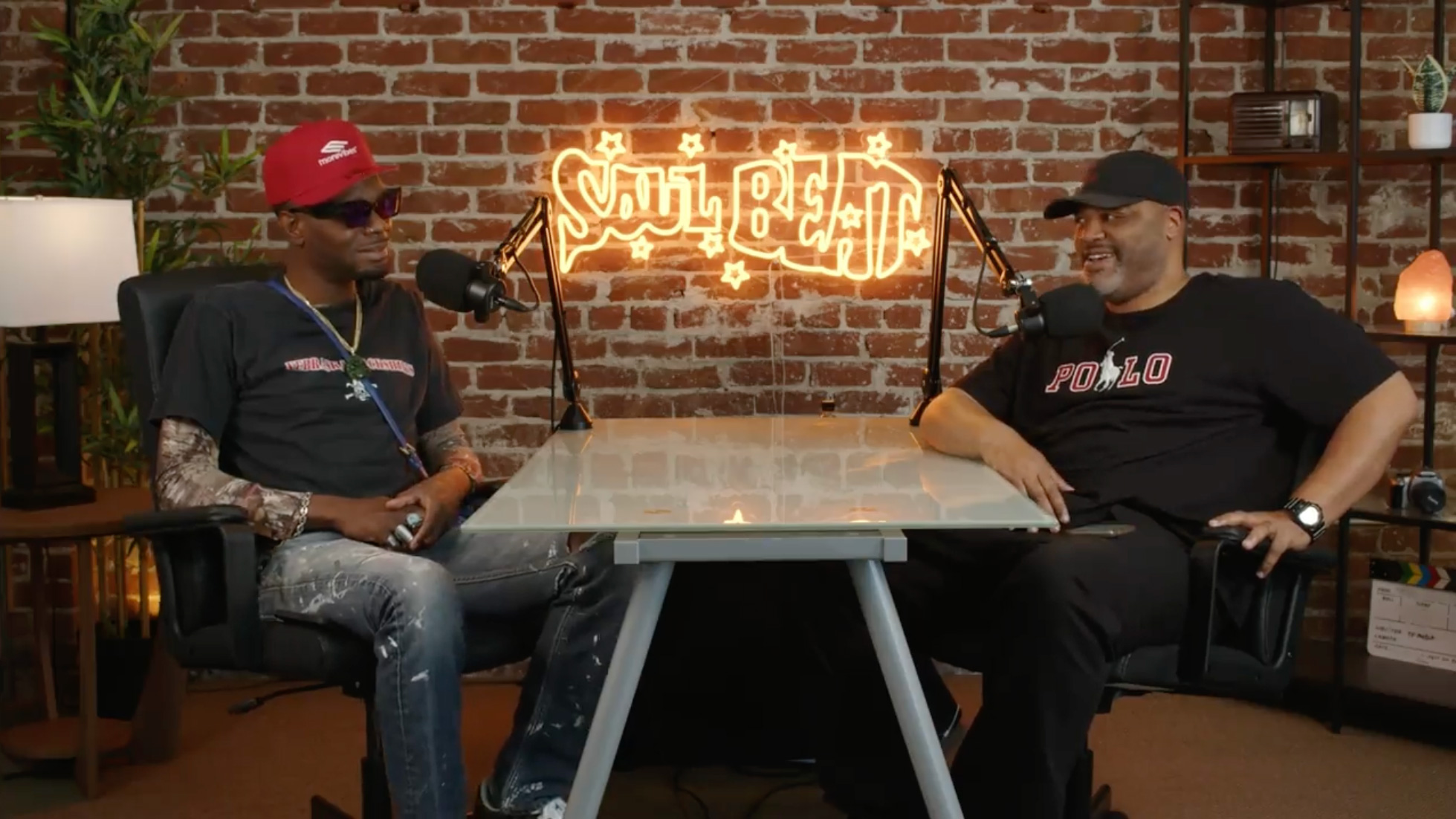Two Black men sit at a table facing each other, with microphones, while a yellow neon sign reading Soul Beat hangs on a brick wall behind them