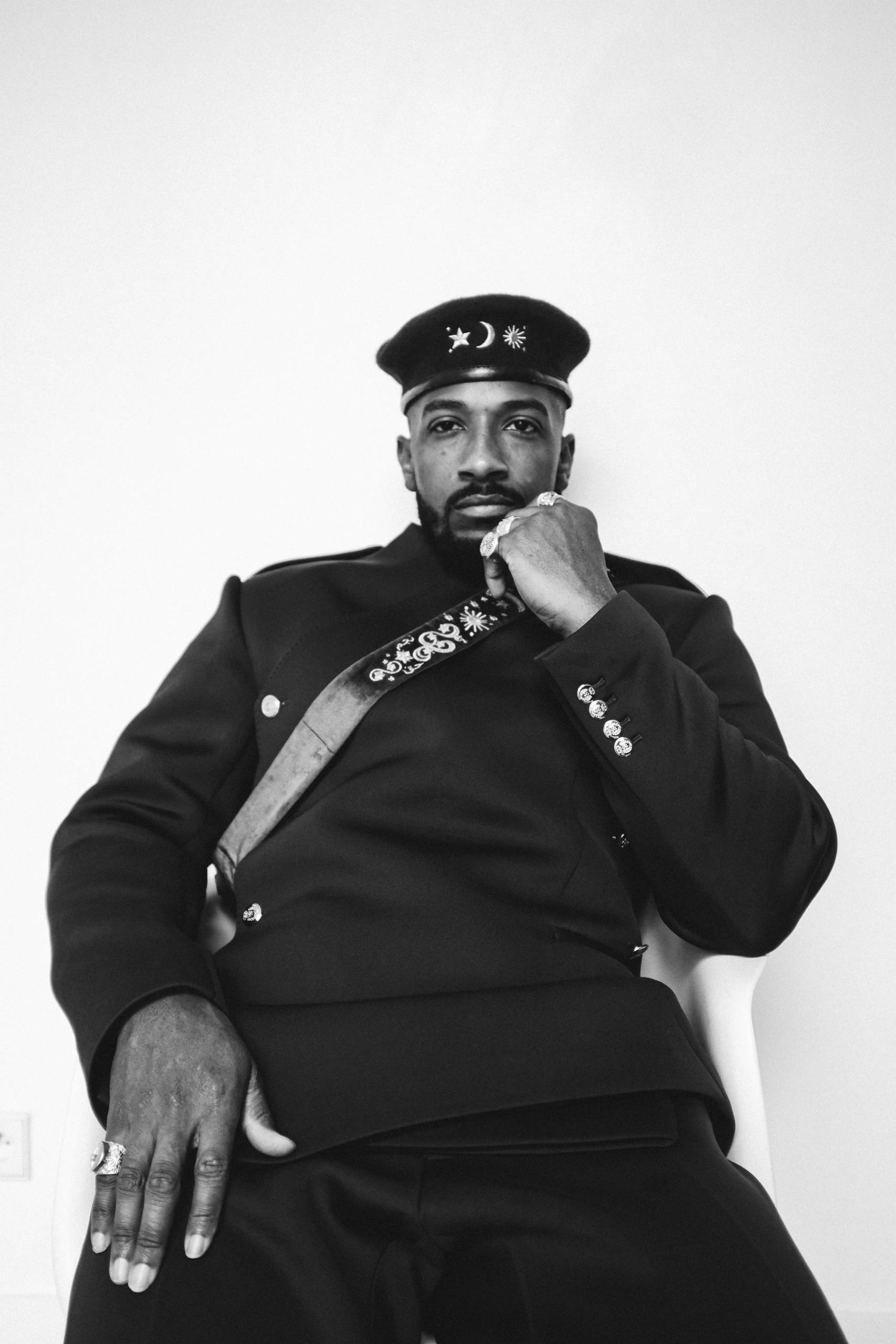 A black and white photo of a man sitting with a hat and jacket on and his left fist to his chin.