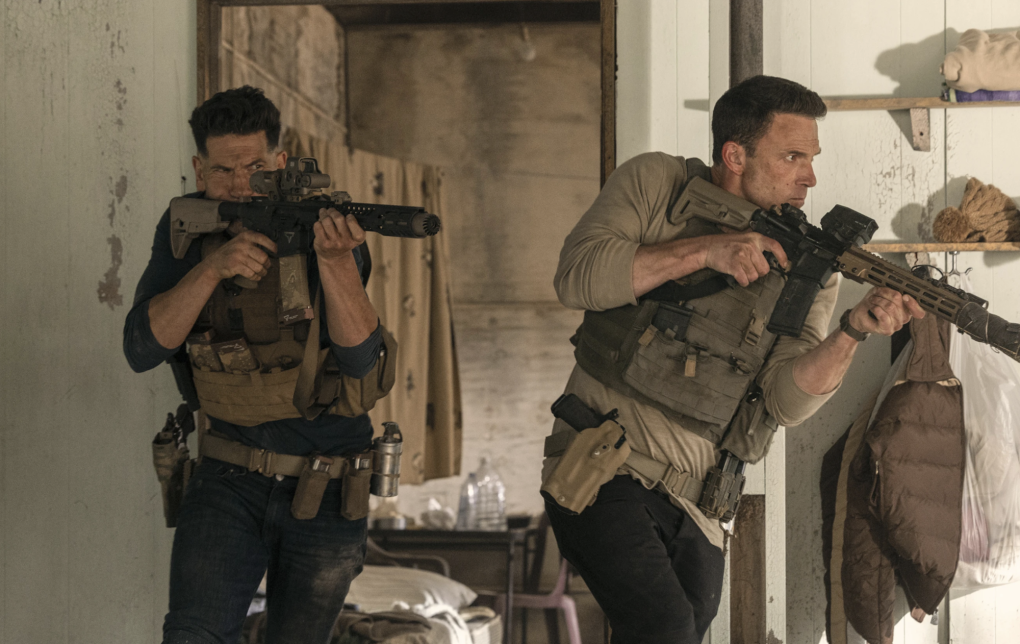 Two men wearing bullet proof vests rush through a door holding up large rifles. They are both dusty.