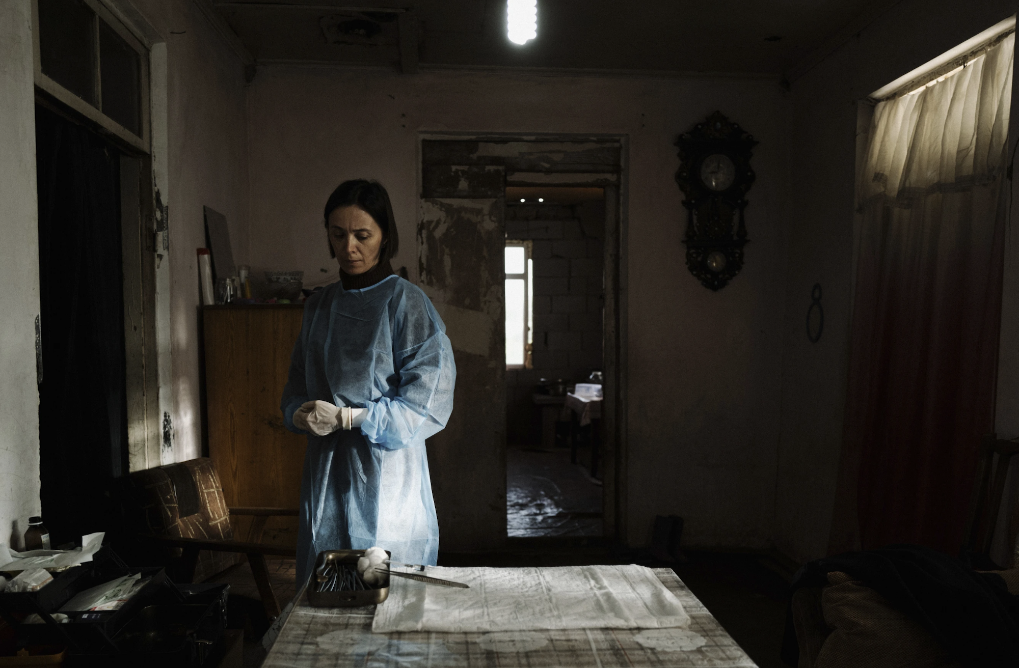 A woman in medical scrubs and gloves stands in a kitchen looking grimly at a tray of surgical instruments.