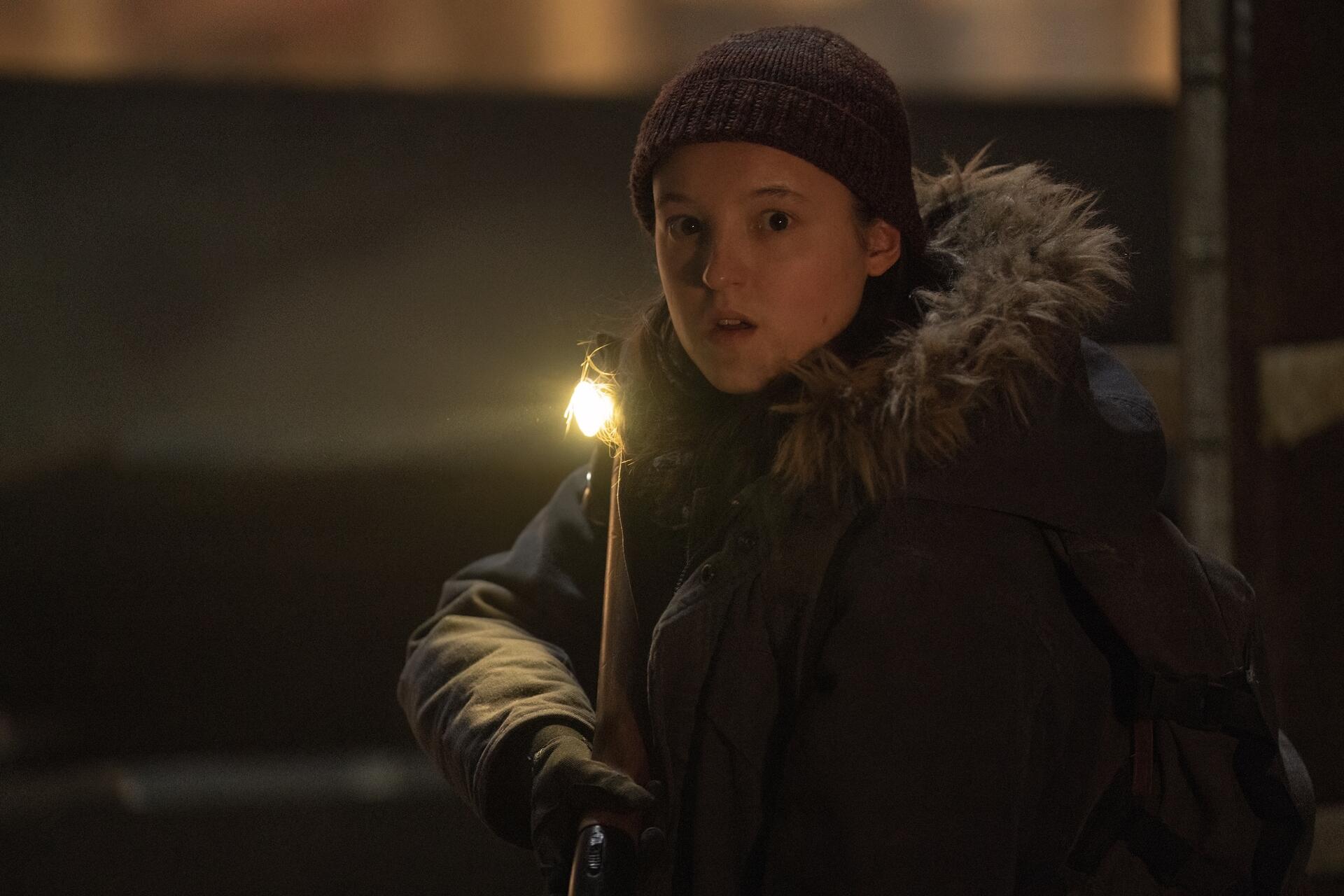 A white adolescent girl wearing a winter coat and beanie stands with a small light in the dark, looking worried.