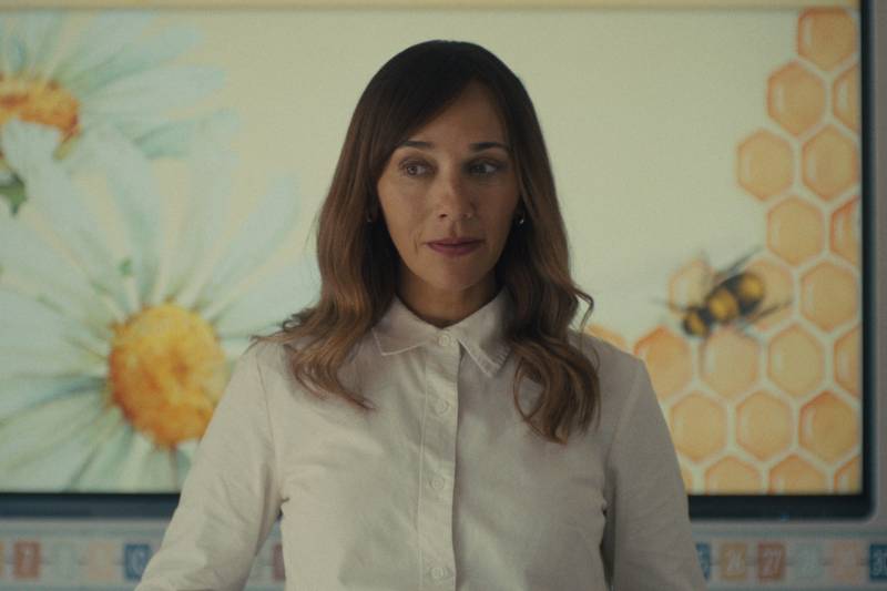 A mixed race woman with long brown hair stands in front of a large screen showing illustrated bees and honey. She is wearing a white button-up shirt and looks uncertain.