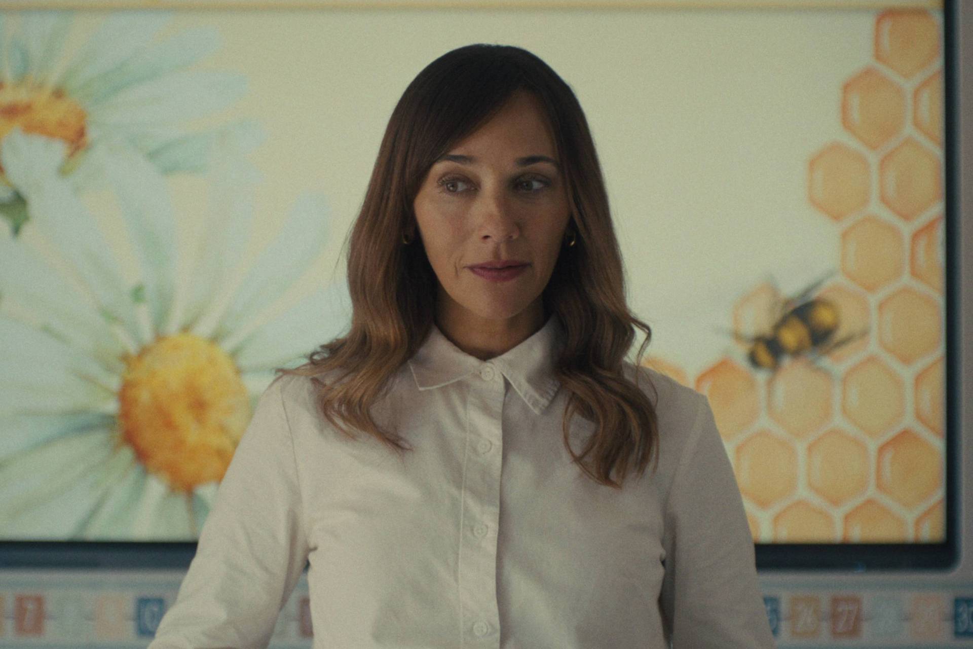 A mixed race woman with long brown hair stands in front of a large screen showing illustrated bees and honey. She is wearing a white button-up shirt and looks uncertain.