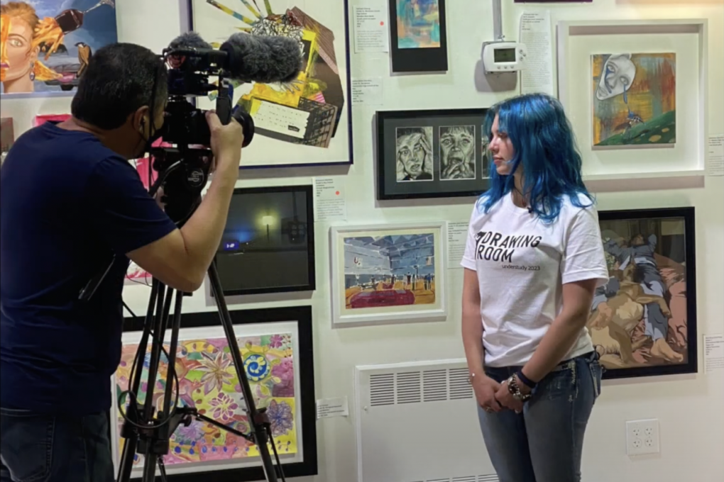 A teenage girl with blue hair wearing a white t-shirt and black jeans stands next a wall of artwork while a cameraman films her.