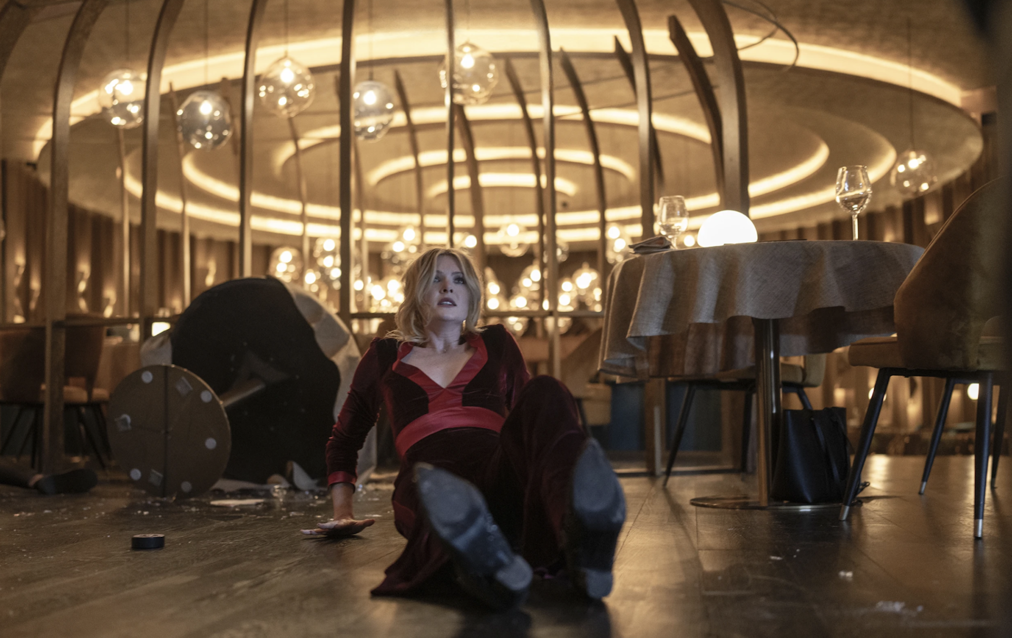 A blonde white woman wearing a dark red evening dress and black boots looks up from the floor of a well-lit fancy restaurant. She is sitting slightly up and appears to be alone.