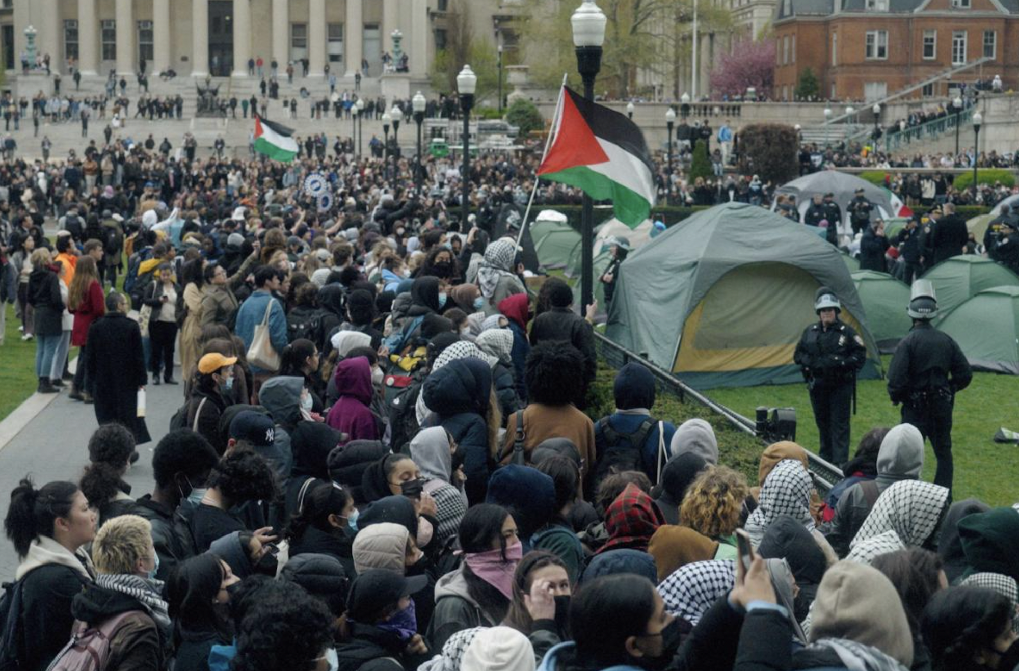 Large numbers of people gather outside on a university campus. Some are holding Palestinian flags. Erected tents are visible on the right.