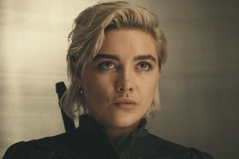 Close up of an attractive young white woman with her blonde hair pulled back and a little disheveled. She is dressed in a black jump suit and carrying a weapon on her back.