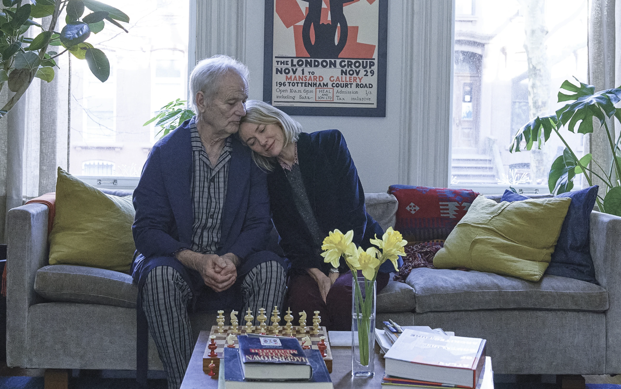 A blonde woman sits on a couch in a well lit, comfortable apartment, leaning her head on the shoulder of an older man who's wearing pajamas.