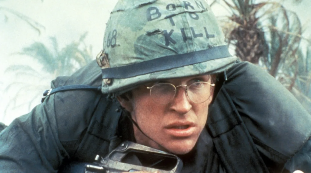 A white man wearing round spectacles crouches low. He is wearing a green army helmet with the words BORN TO KILL written on it.