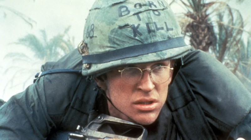 A white man wearing round spectacles crouches low. He is wearing a green army helmet with the words BORN TO KILL written on it.