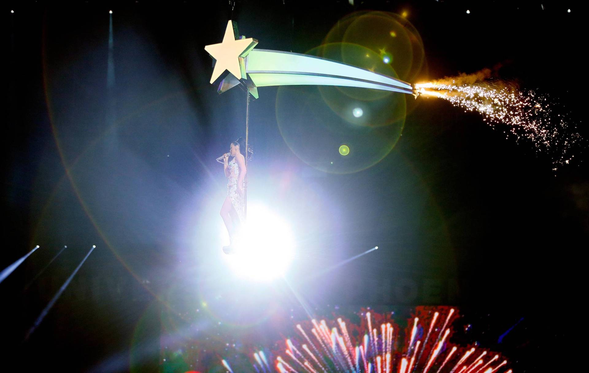 A woman attached to a shooting star prop soars over an audience, fireworks exploding in the background.