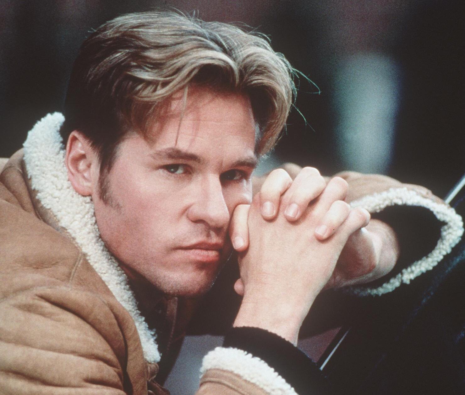 A white man wearing a sheepskin coat leans on his hands which are clasped together.
