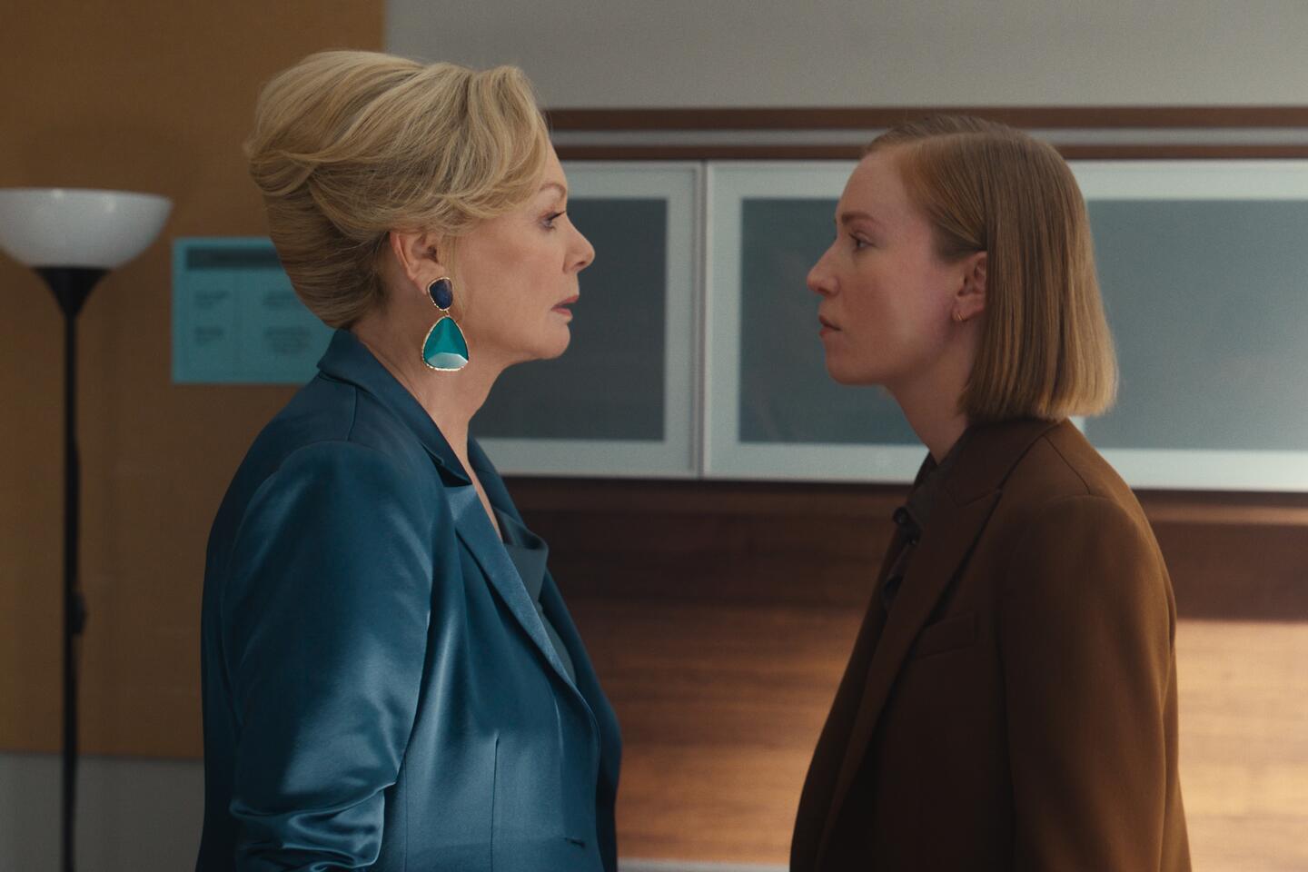 Two white women - one older, wearing a satin blue suit, one in her late 20s, wearing a brown jacket - stand sourly, face to face, inside an office.