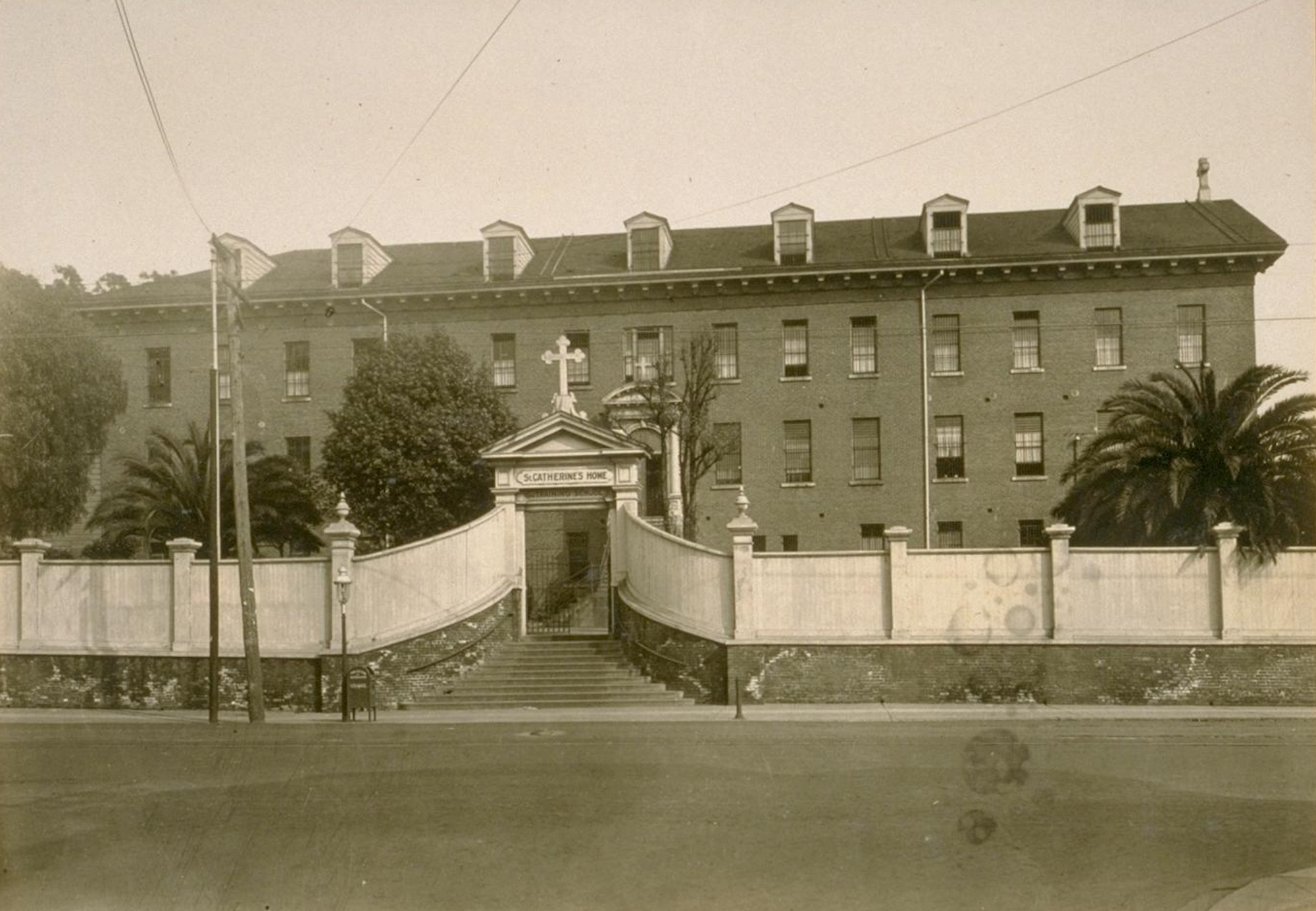 A sepiatoned archival photo of a hotel-like building, with a brick facade and many windows, surrounded by a white fence and large gate adorned with a cross.