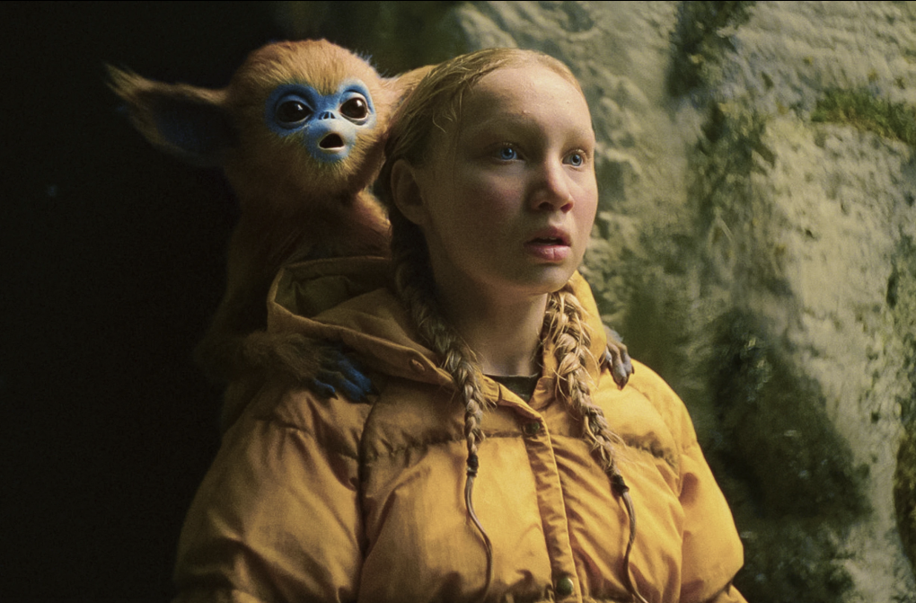 A young girl with blonde braids and a yellow anorak stands next to a cave with a small, cute magical, creature on her shoulder.
