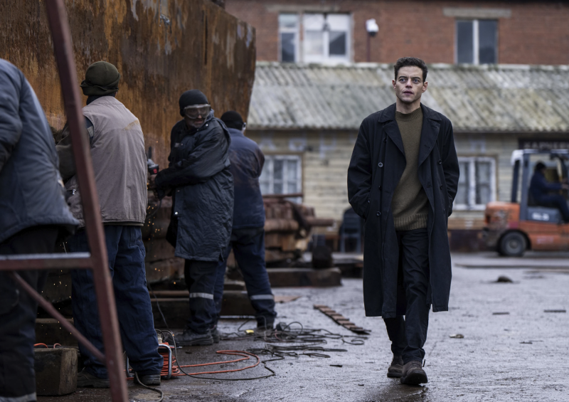A mysterious man wearing a long black coat walks down a squalid street.