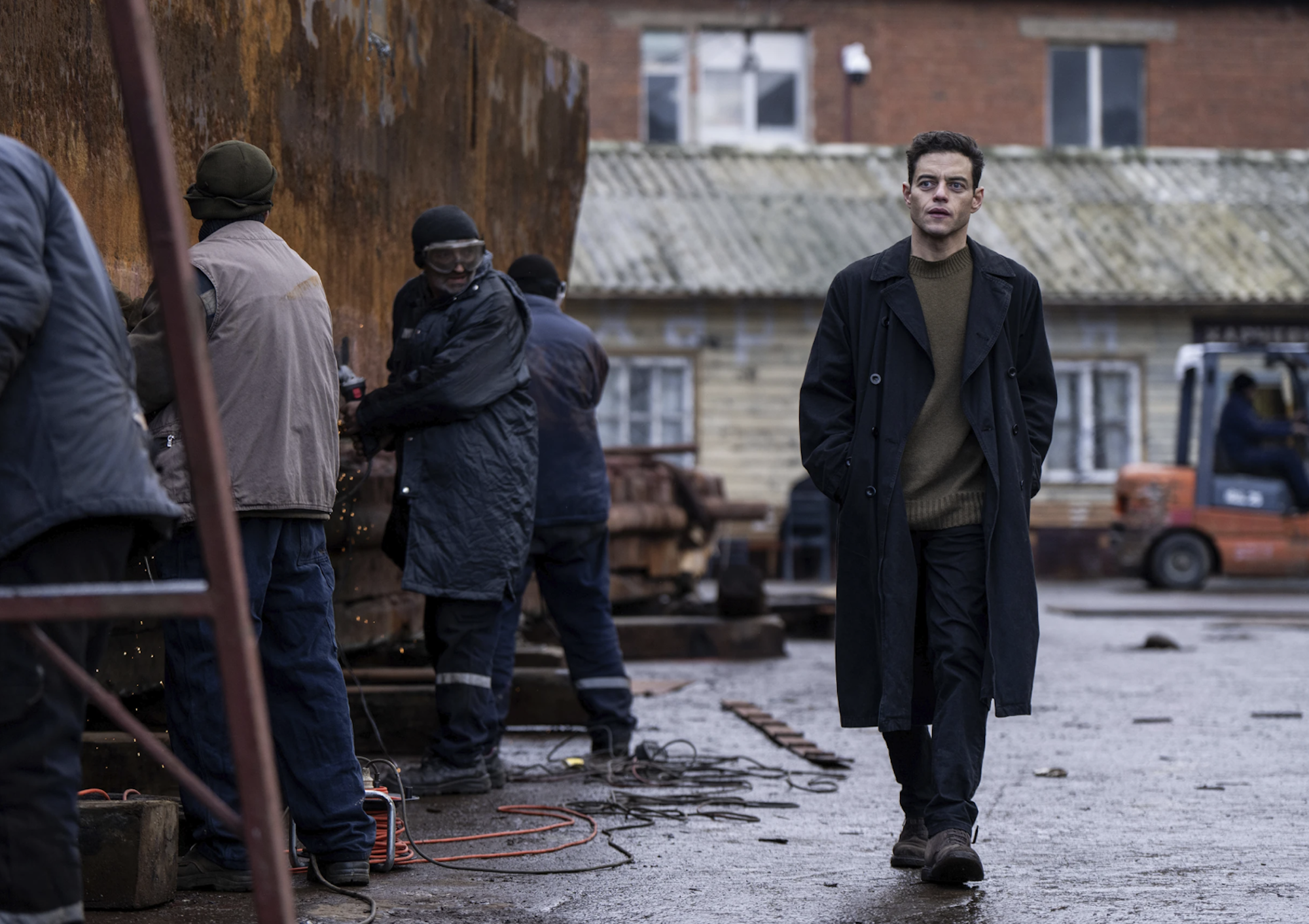A mysterious man wearing a long black coat walks down a squalid street.