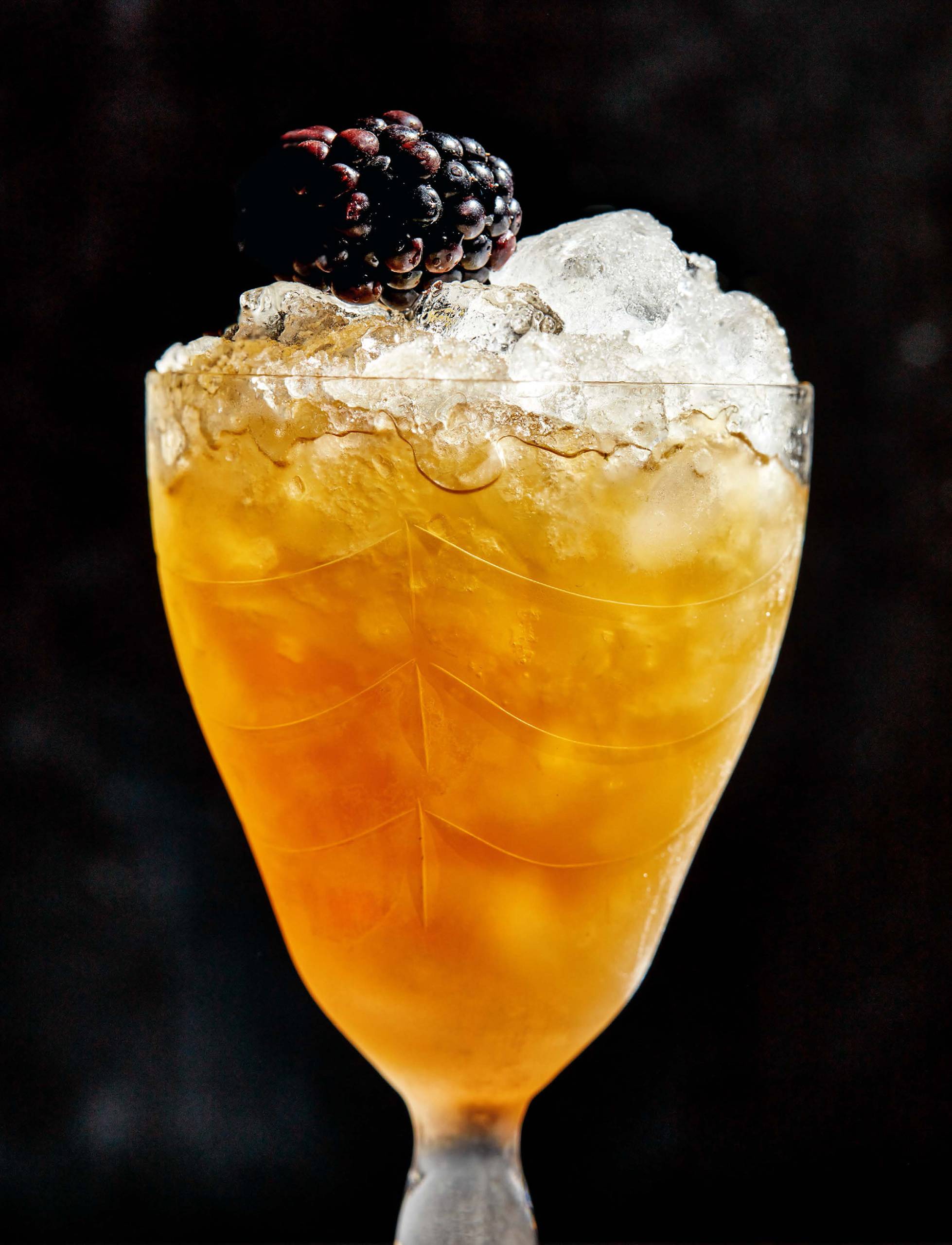 Amber-colored cocktail with ice, garnished by a blackberry, in a sherry glass.