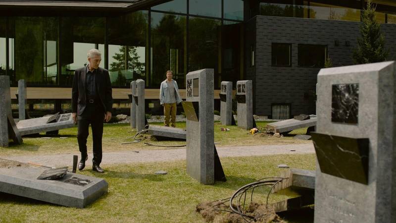 A man stands in a modern graveyard, surrounded by tombstones containing video screens. A woman stands in the distance.