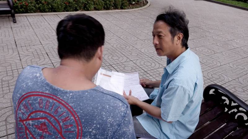 An over-the-shoulder shot of two men talking while sitting on a park bench.
