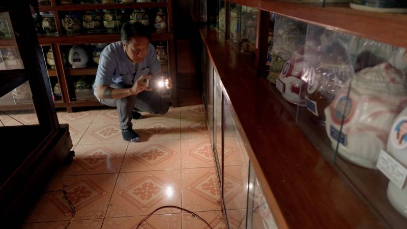 A man crouches while holding a cellphone with the light on, looking at a room full of urns. 