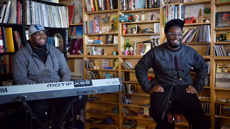 A Black man wearing a black sweater, pants, baseball cap and clear spectacles sits on a stool behind a microphone smiling. At his side is a Black man behind a keyboard, also smiling. They are both seated in front of packed bookshelves.