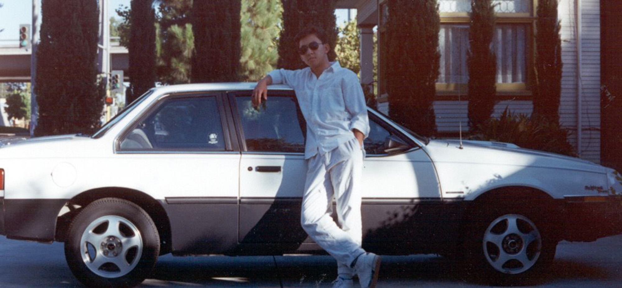 Asian American teenager leans against a sporty white coupe in a photo taken during the 1980s.