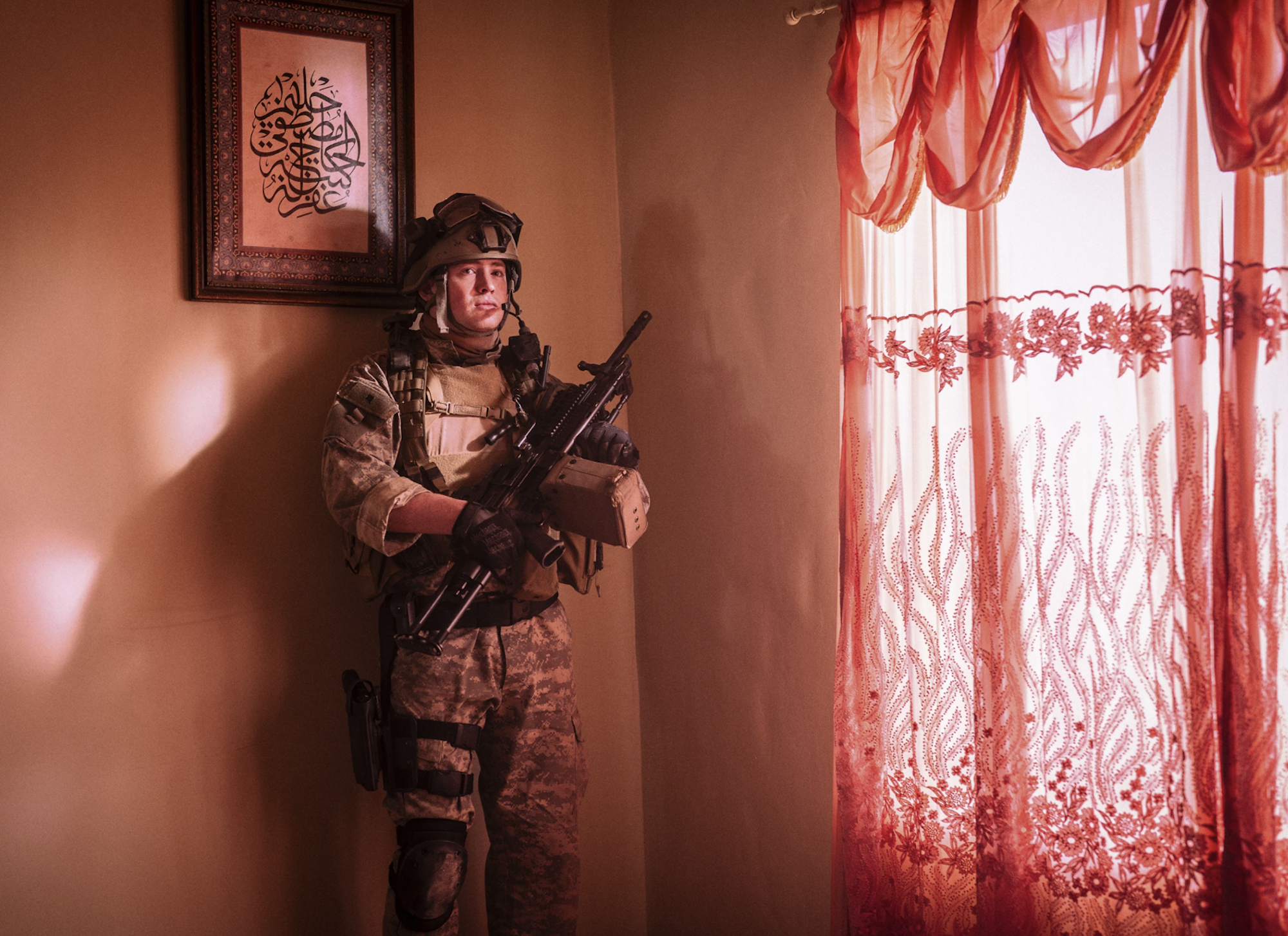 A marine stands up straight, visibly nervous, in the corner of a domestic home. An Arabic piece of art hangs above his helmet. Nearby is a window with transparent red curtains.