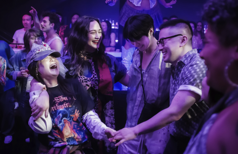A group of friends stand in a circle laughing, inside a purple-lit nightclub. Three of them are Asian, one is Indigenous.