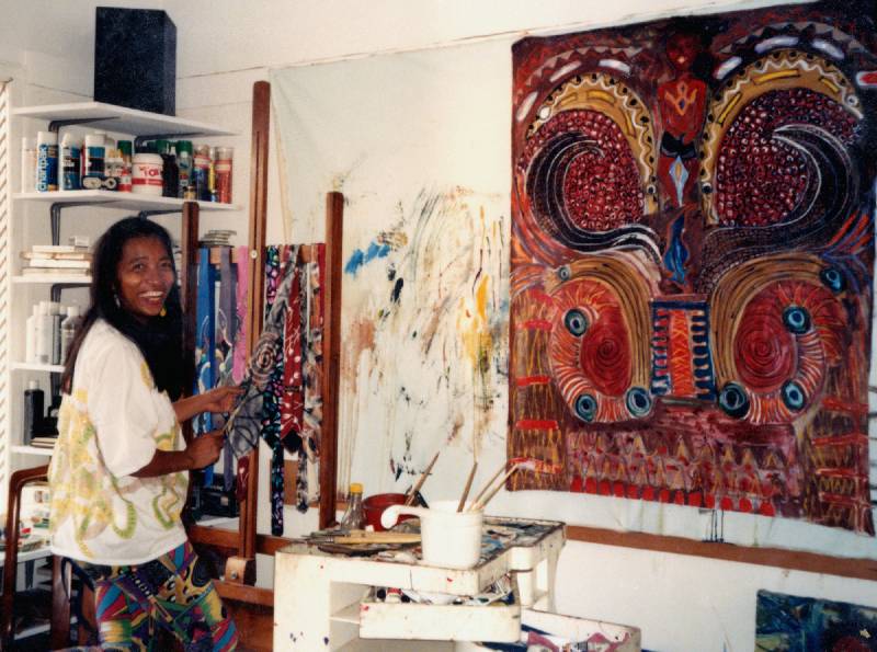 smiling woman with long hair hold paintbrushes in front of large abstract canvas