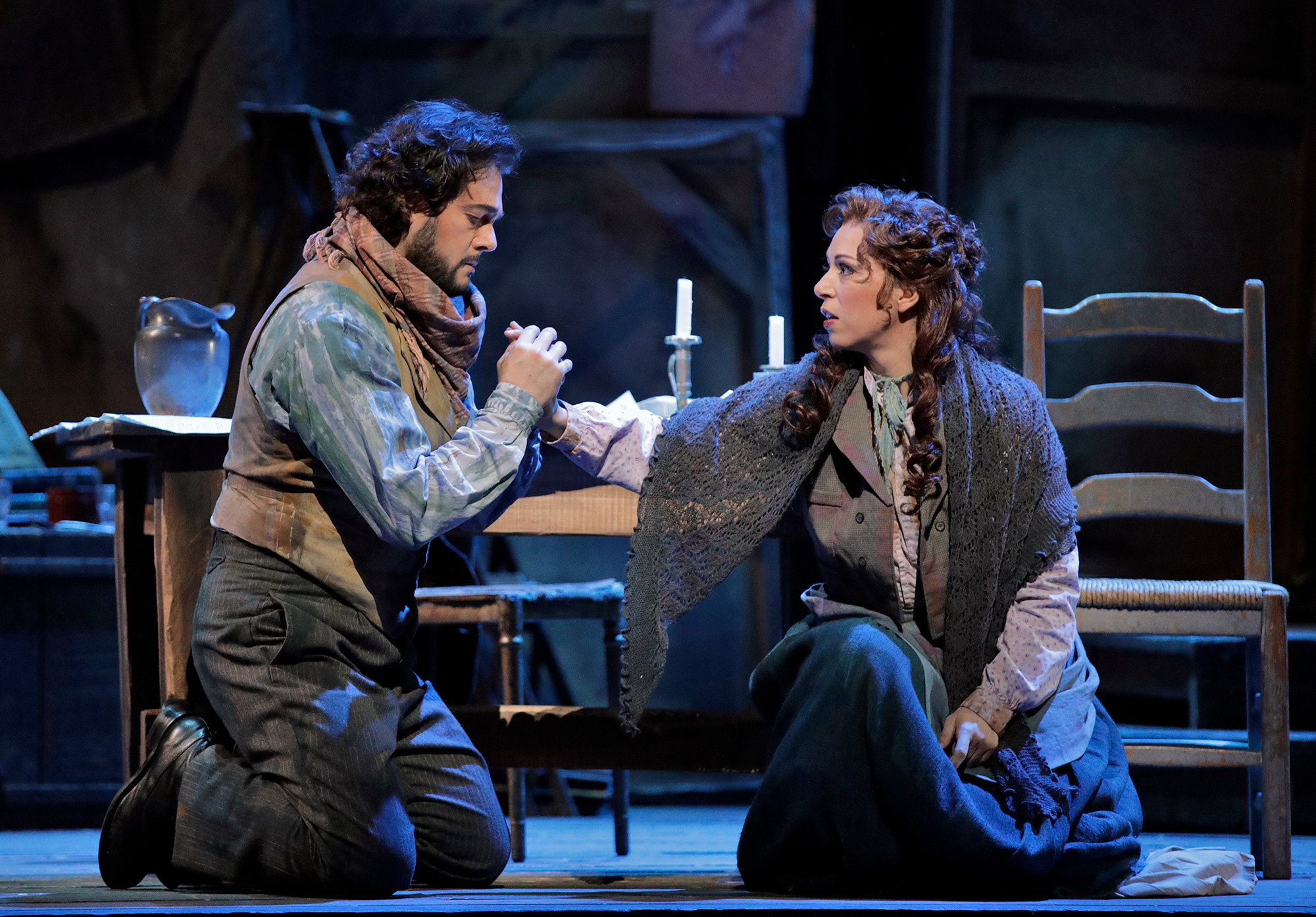 A man in a scarf and ragged clothing holds the hand of a woman, similarly dressed, both kneeling on the ground