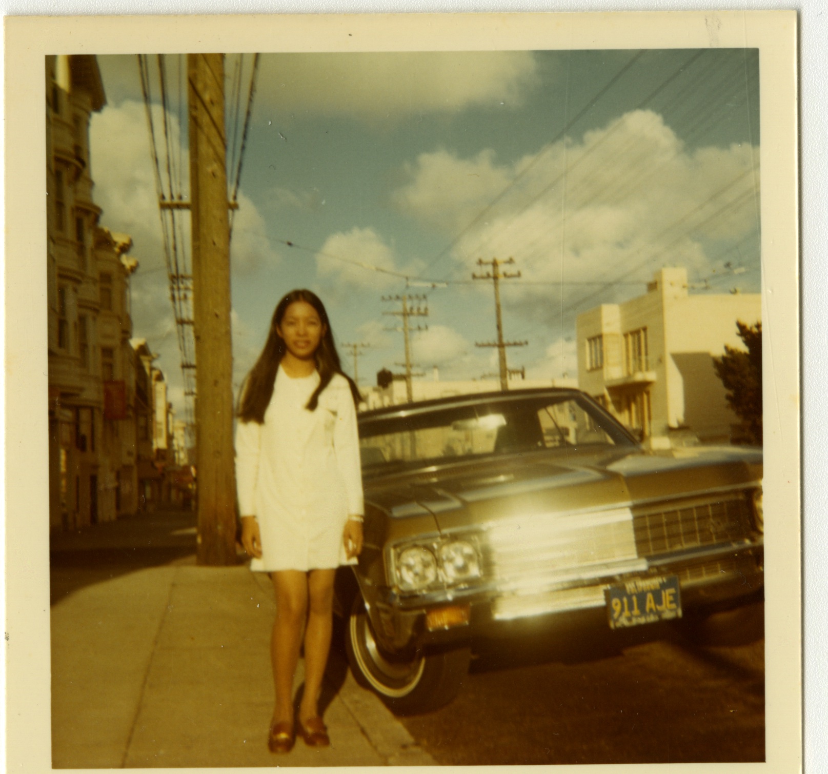 woman with long hair in short white dress next to a car on suburban street