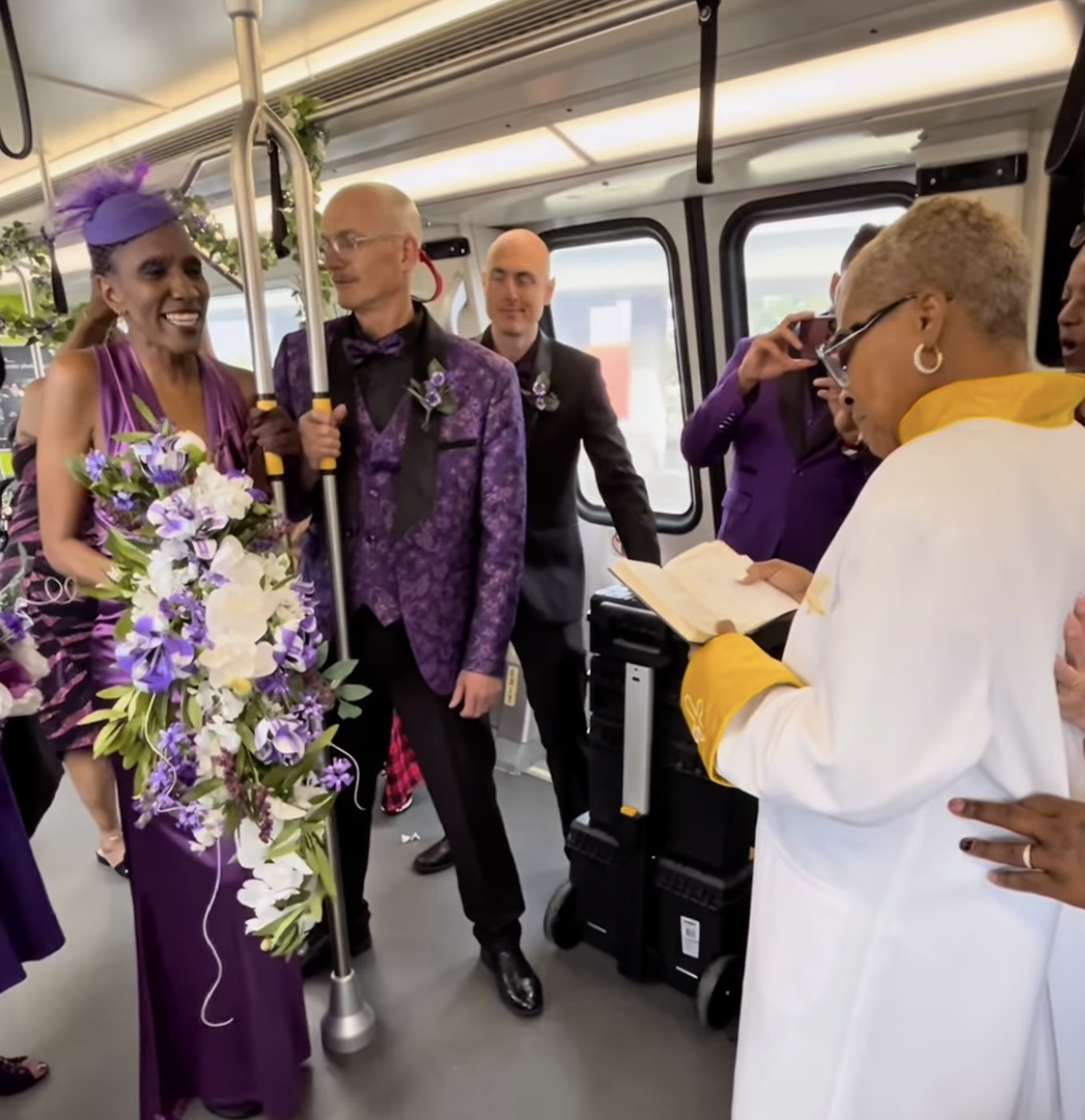 Inside a train carriage, a Black woman wearing a purple satin gown and holding a large bouquet of purple and white paper flowers, stands smiling next to a white bald man in a purple and black suit. Before them is a female pastor in white and yellow robes reading from a small book.