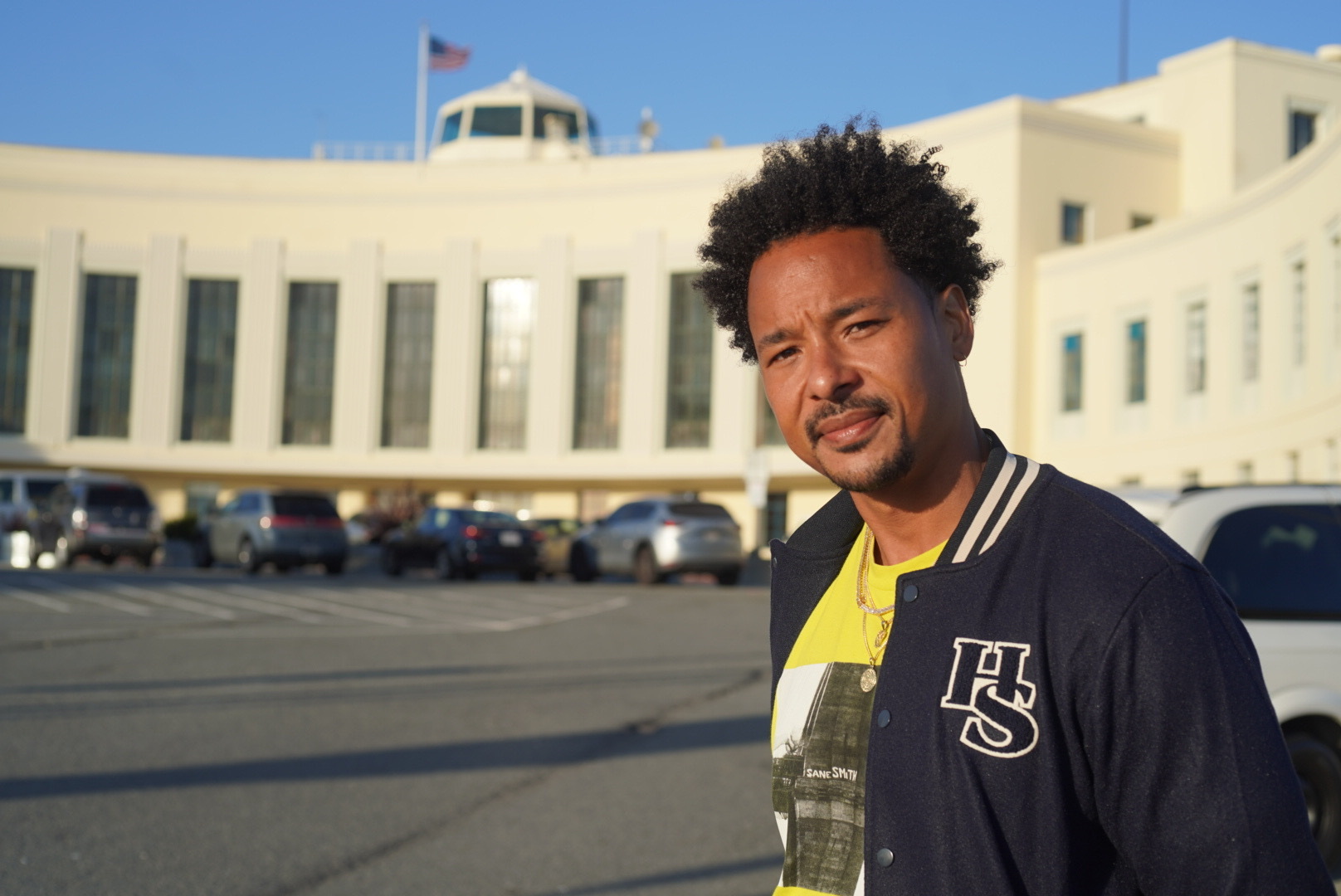 A man in a blue jacket and yellow shirt poses for a photo on a sunny day.