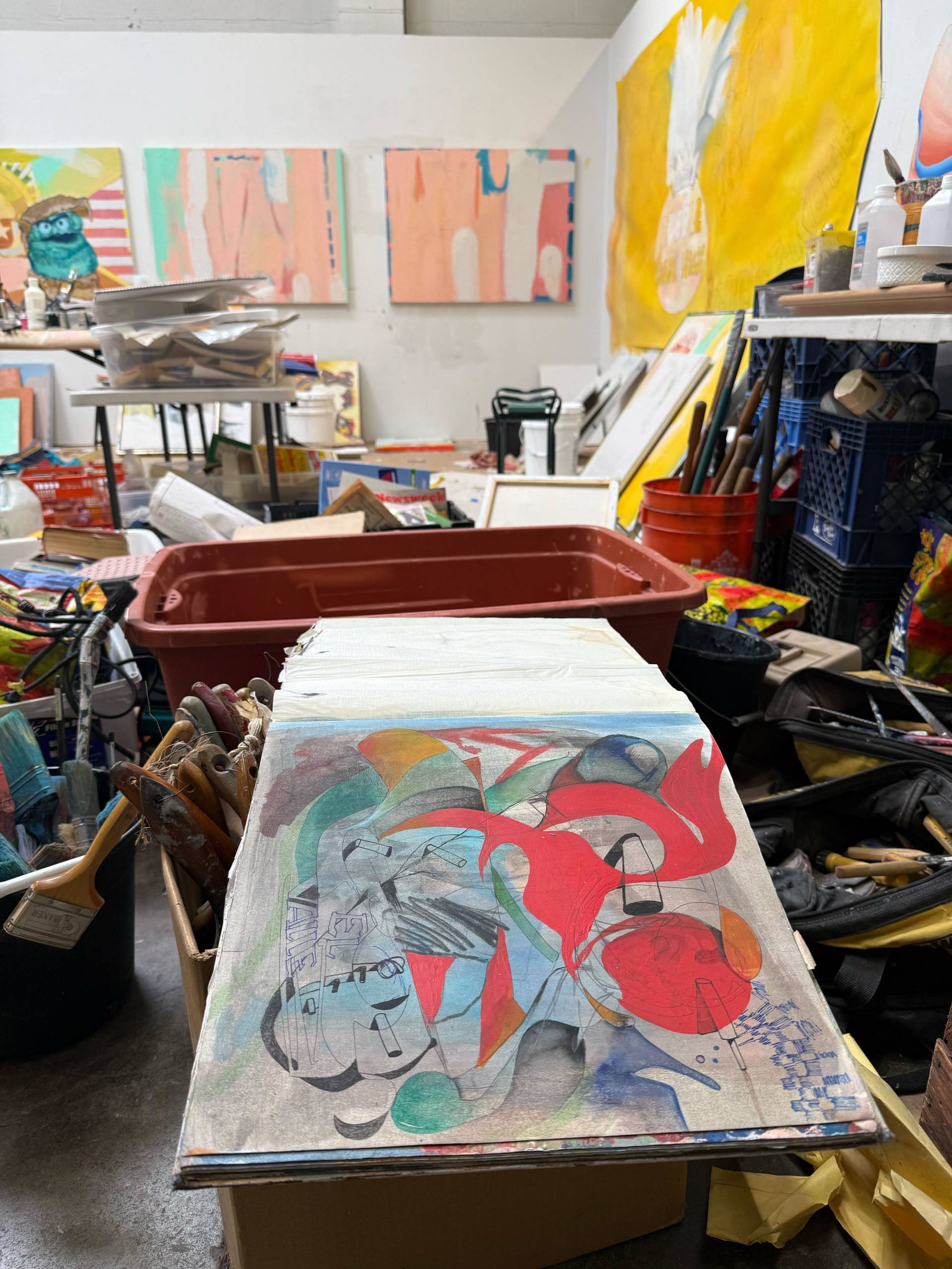 An opened page of a sketchbook reveals a colorful illustration; the book is surrounded by a long list of formerly discarded items in a studio.