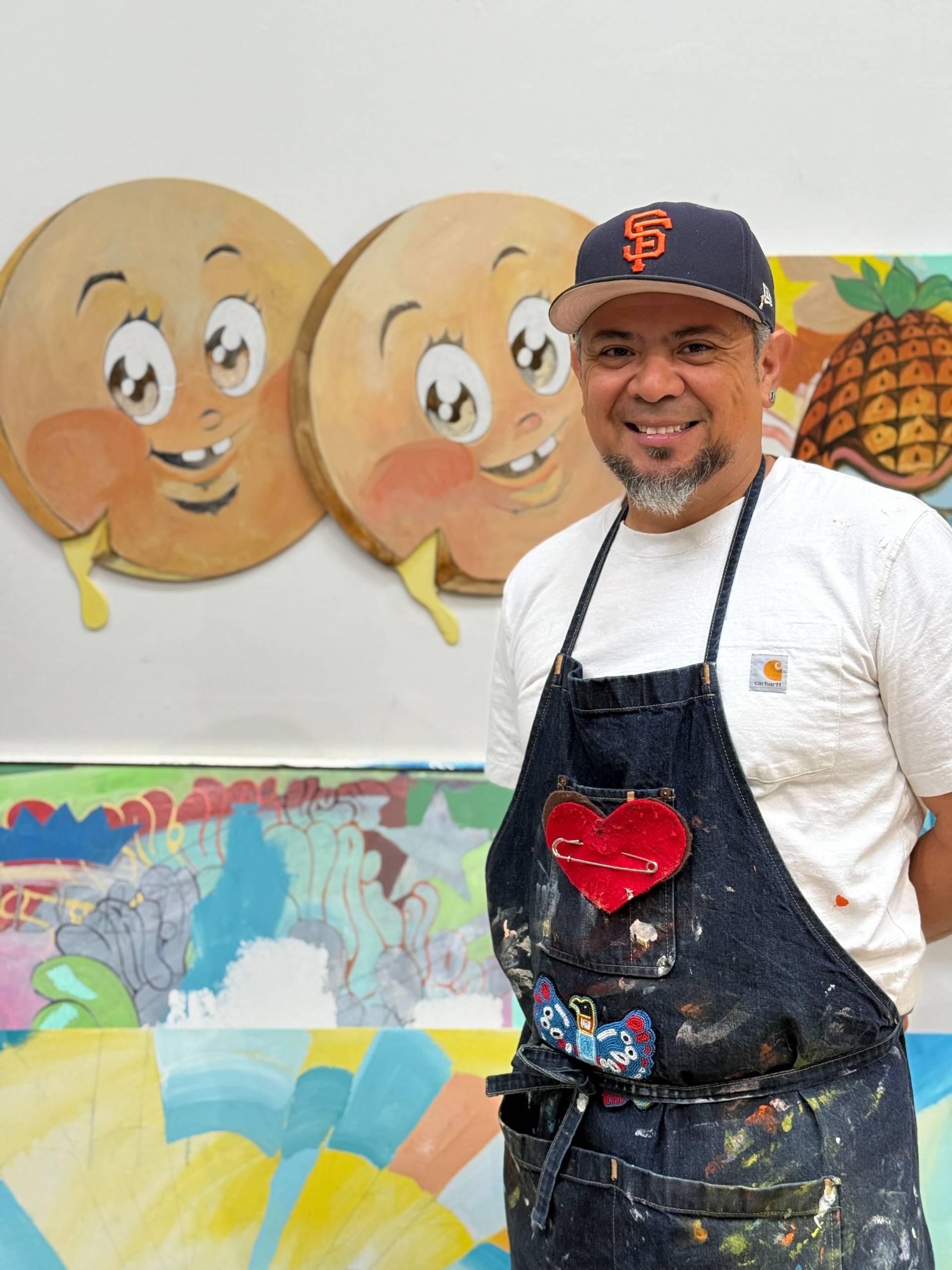 A man with a San Francisco Giants hat and a paint-covered an apron pose for a photo in front of some artwork he's created.