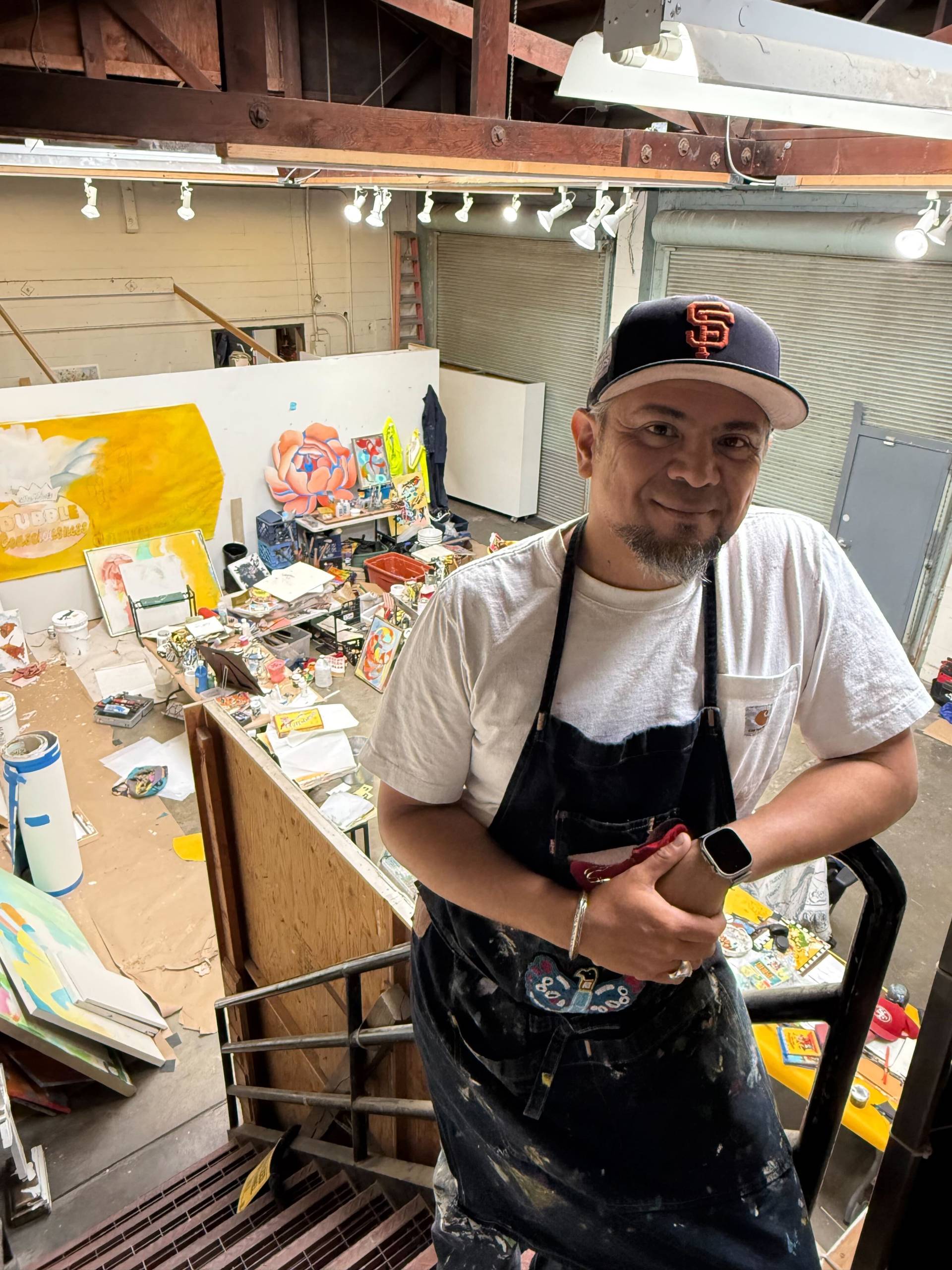 A man with a hat and a paint-covered apron on stands and poses while looking at the camera.