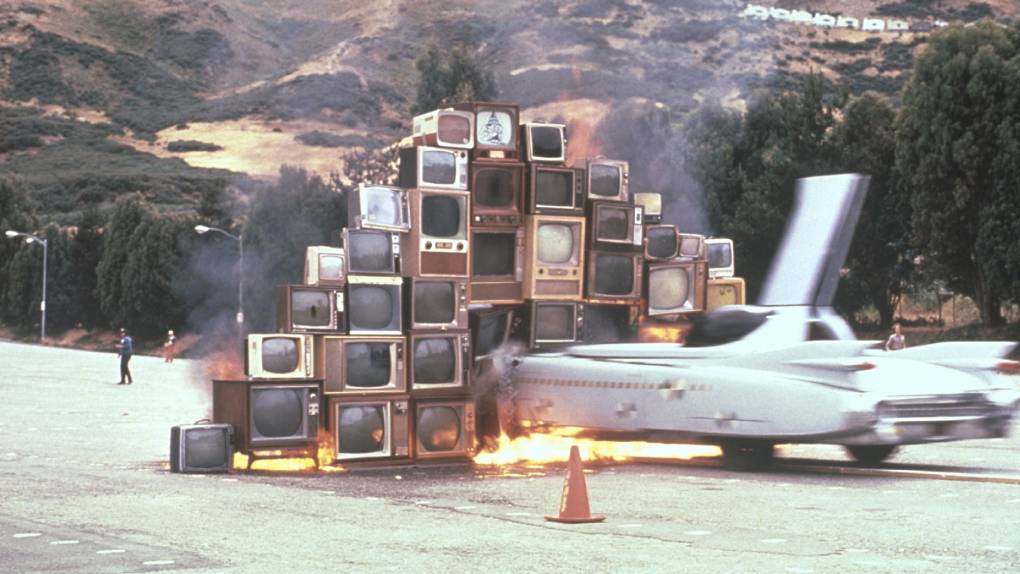 car with large tail fin runs into pyramid of TV sets, flames below