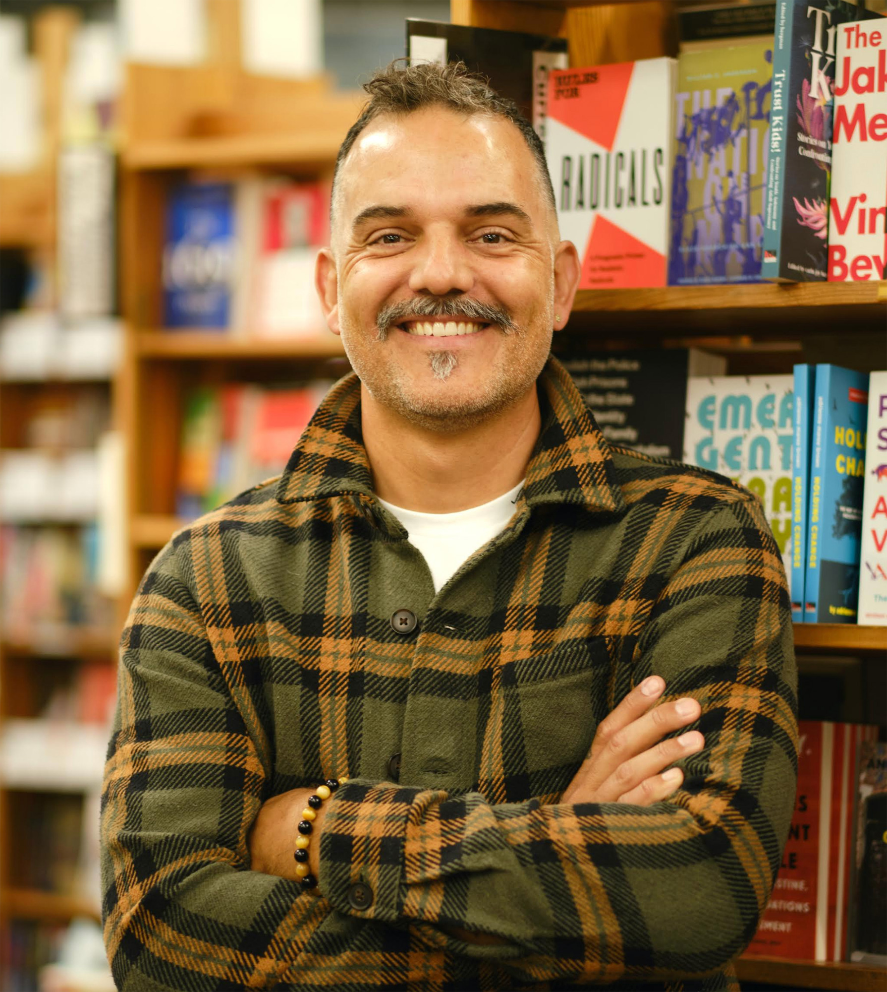 A smiling man in a plaid flannel with folded arms against a backdrop of bookshelves