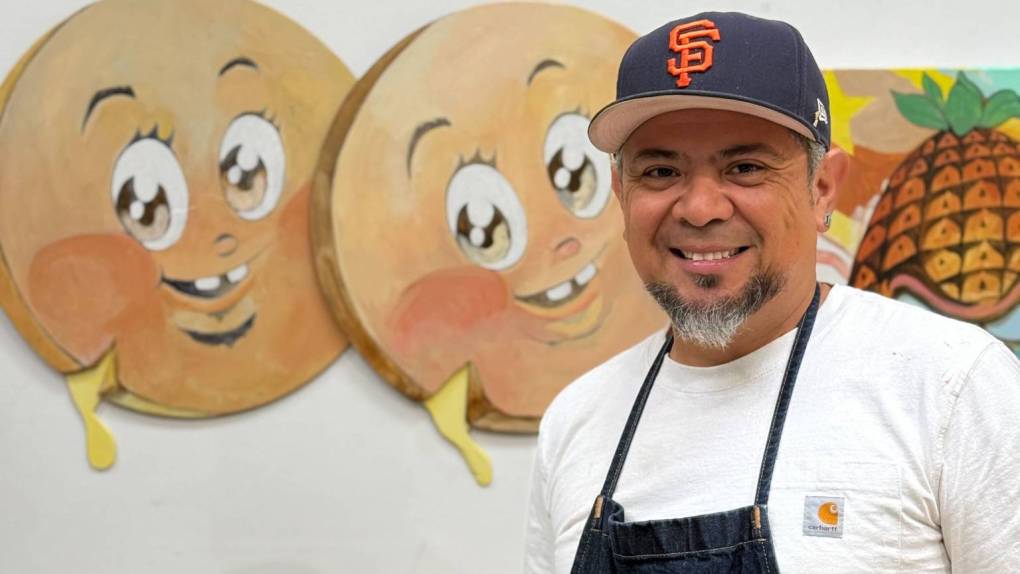 A man in a SF Giants hat and overalls with a salt-and-papper beard stands in front of a wall with circular face characters made of wood