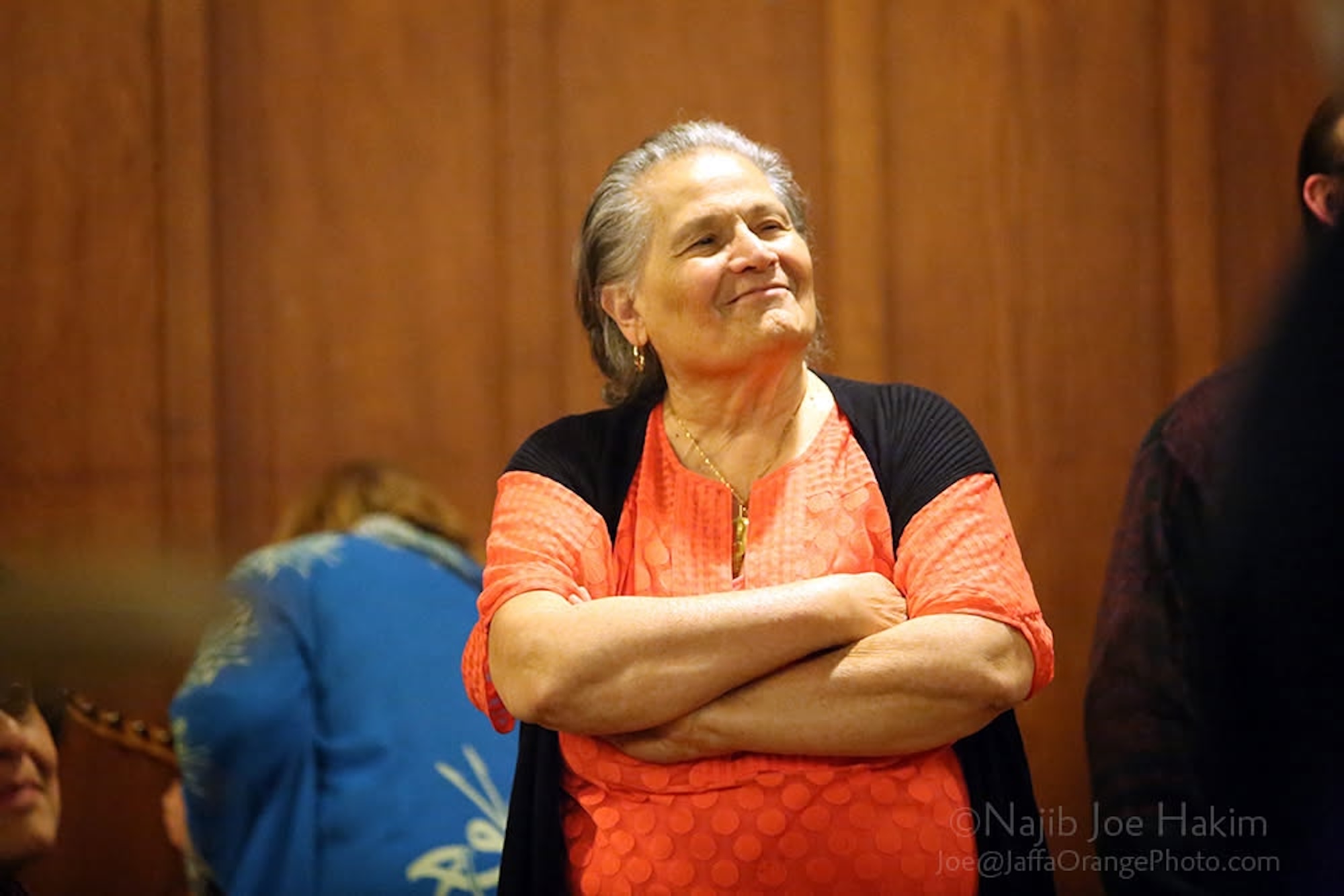 A senior woman with grey hair smiles, chin slightly lifted, with her arms folded in front of her. She is standing in front of a wood paneled wall.