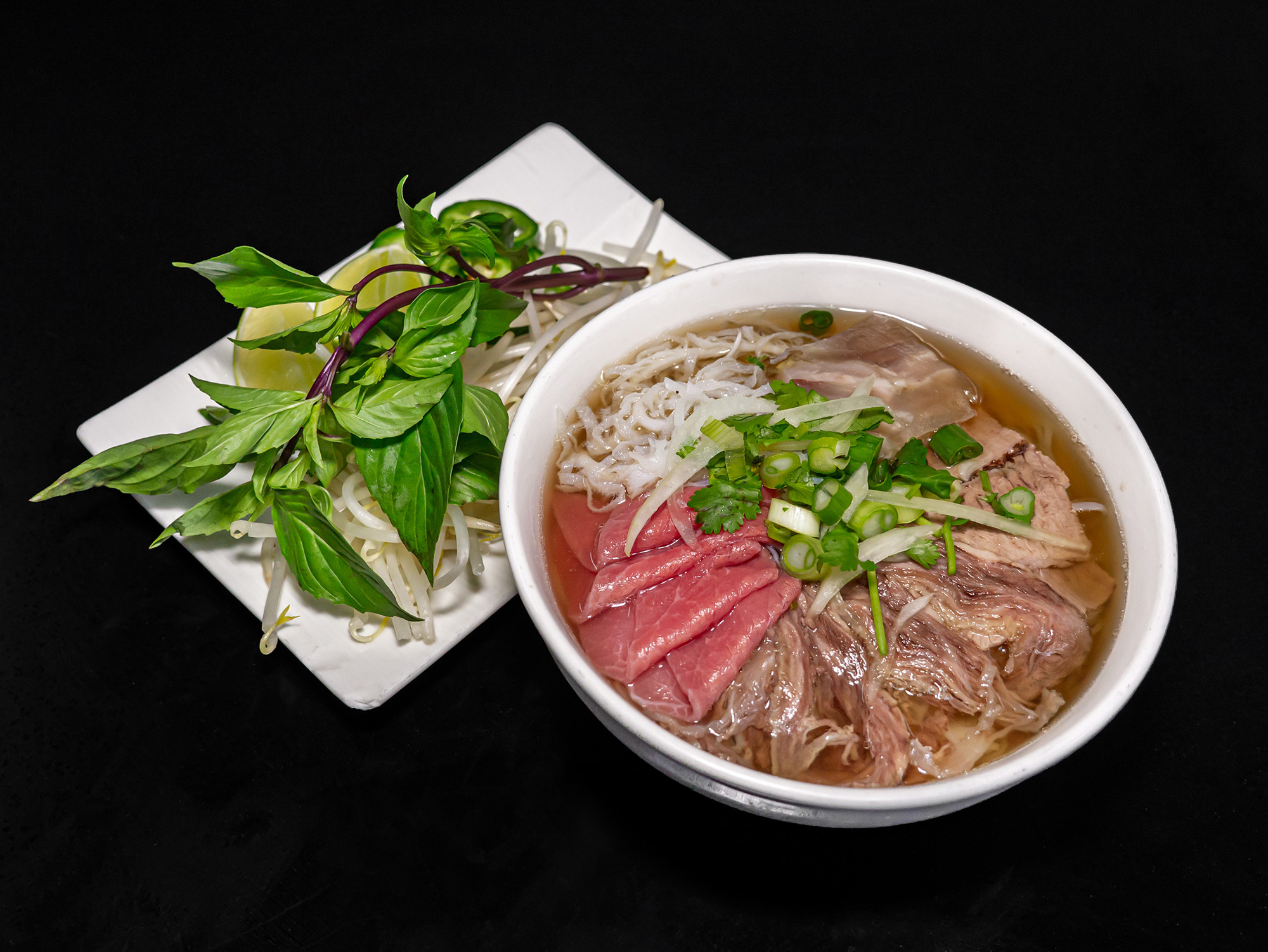 Bowl of pho with accompanying herbs against a black background.