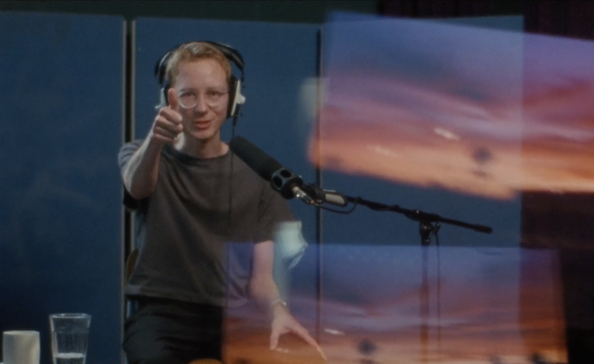 A thin white man with red hair talking in a sound booth while wearing headphones. 