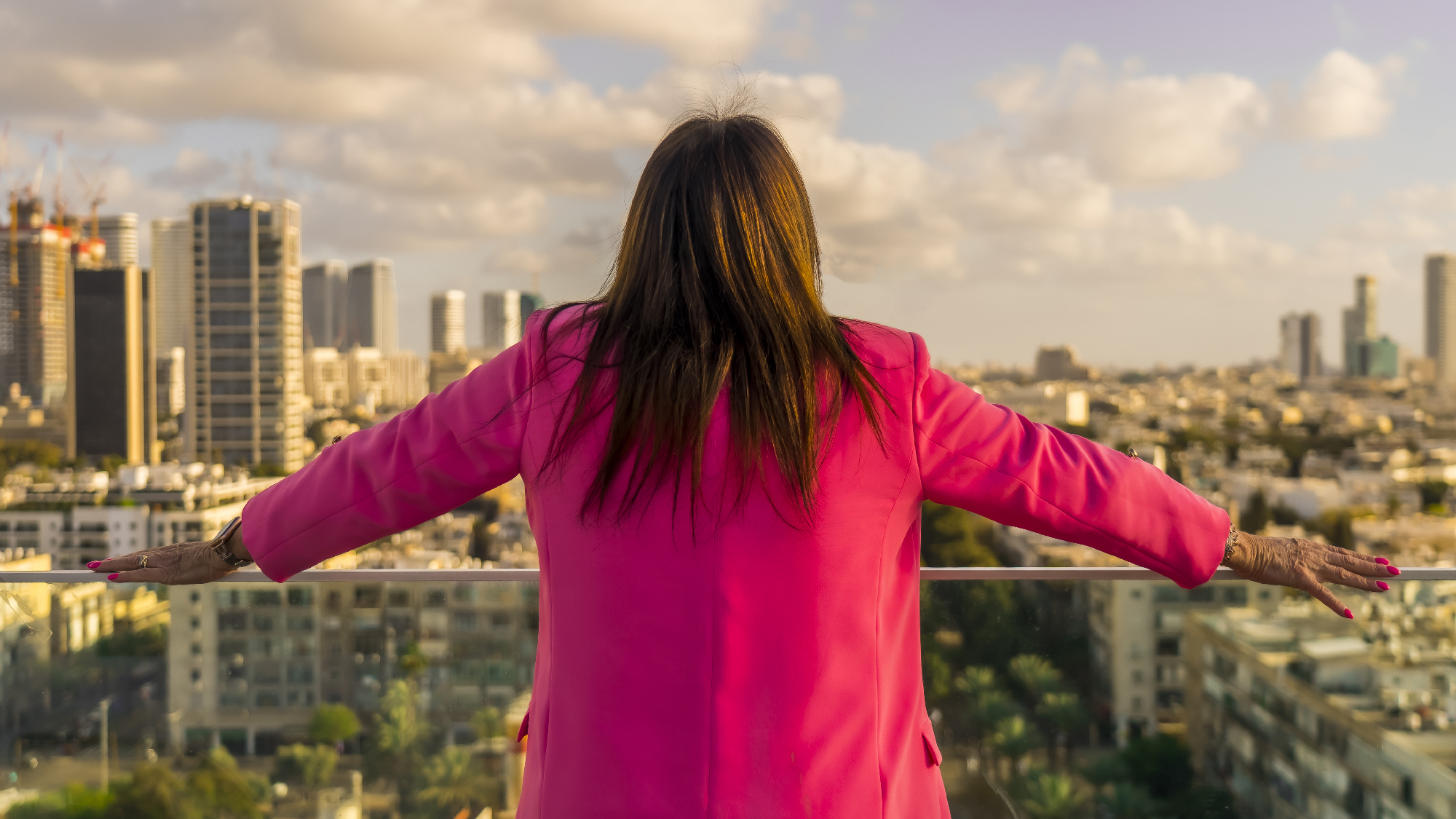 person with long brown hair in bright pink blazer looks over city with back to camera, arms spread