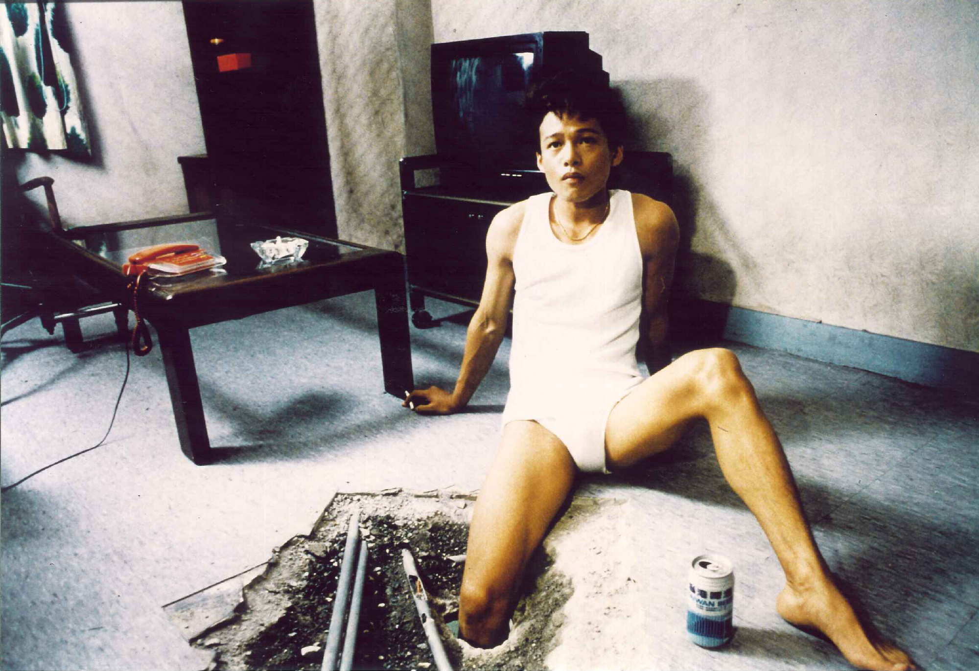 man in underwear and white tank sits on floor with leg through hole, with beer and cigarette
